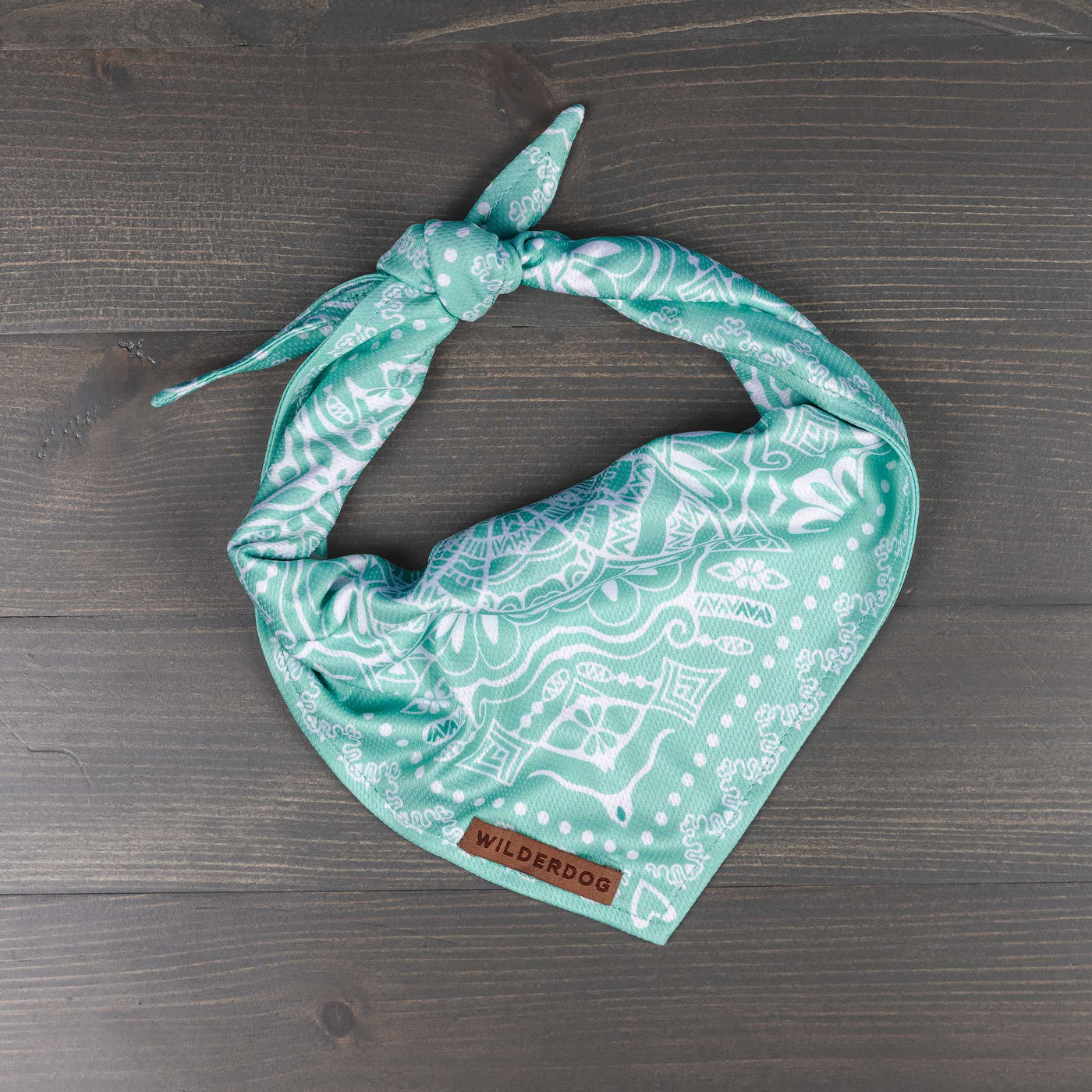 An Agave Bandana, featuring a teal and white pattern with intricate designs and a knot at the top, is laid out on a gray wooden surface. This stylish bandana includes a small leather tag labeled "Wilderdog" near the bottom, making it an ideal addition to your trail dog accessories collection.