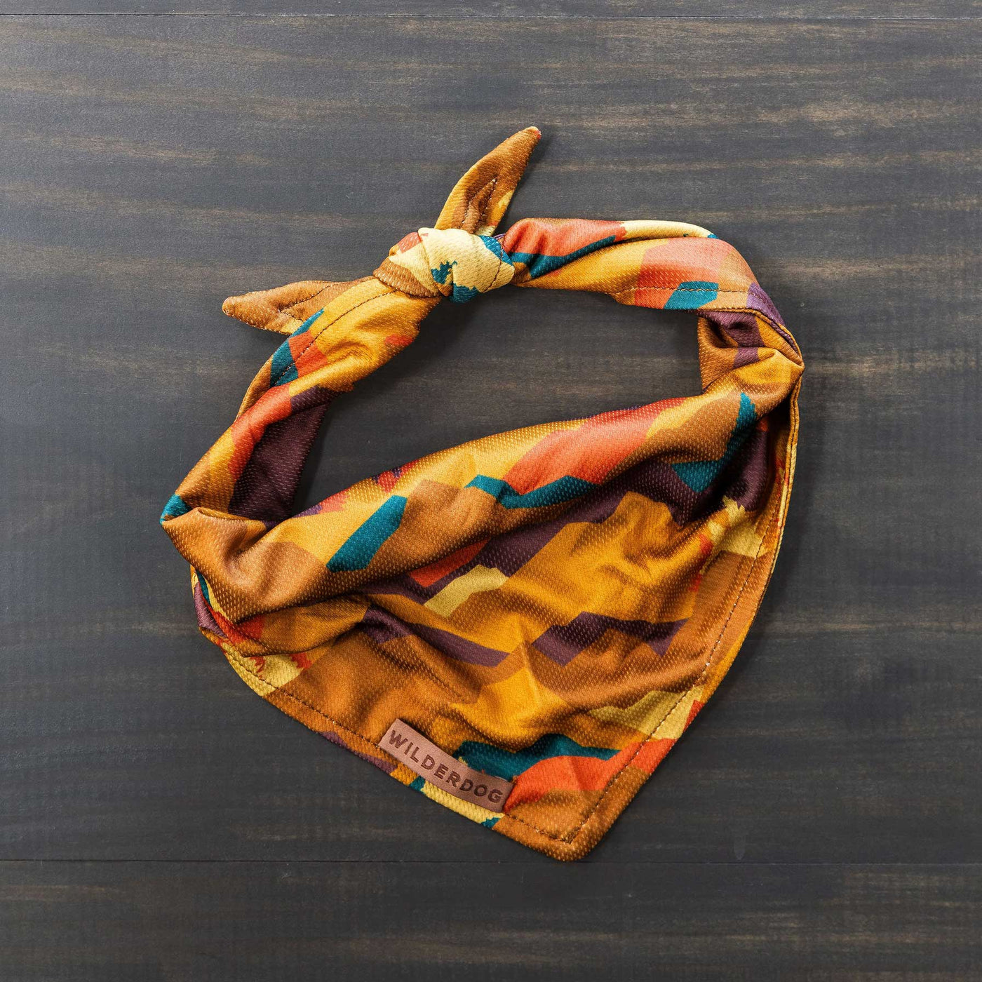 The Autumn Air Bandana, adorned with an abstract pattern in vibrant shades of orange, yellow, and teal, is neatly folded on a dark wooden surface. Perfect as part of your dog's trail gear, a small tag on the bandana reads "Wilderdog.