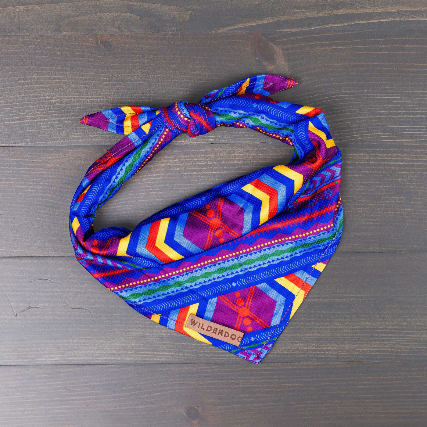 The Big Sky Bandana, featuring a colorful geometric pattern in shades of blue, red, yellow, green, and purple along with a small leather patch bearing the brand name "Wilderdog," lies on a wooden surface. Styled in a folded and knotted arrangement, this vibrant cooling bandana is perfect for trail adventures.