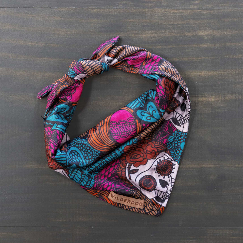 The Calavera Bandana, made from a colorful poly-blend mesh and adorned with a vibrant Day of the Dead (Día de los Muertos) design, is neatly folded on a wooden surface. Featuring intricate patterns of skulls, hearts, and flowers in shades of pink, teal, orange, and white, this cooling bandana is perfect for your dog's trail accessories. A tag reads "Wilderdog.