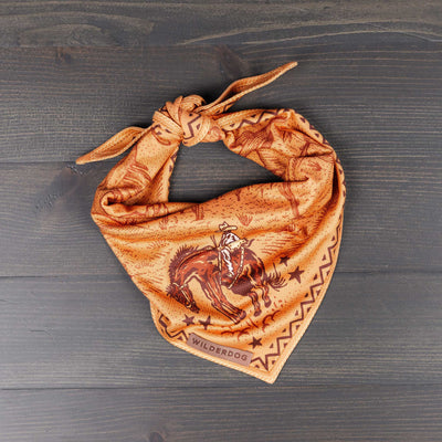 The Cowpoke Bandana is an orange bandana featuring a vintage illustration of a cowboy on horseback, surrounded by stars and decorative patterns. This quick-drying trail dog accessory is neatly folded and tied, with the brand name "Wilderdog" visible in the corner. It is laid on a dark wooden surface.