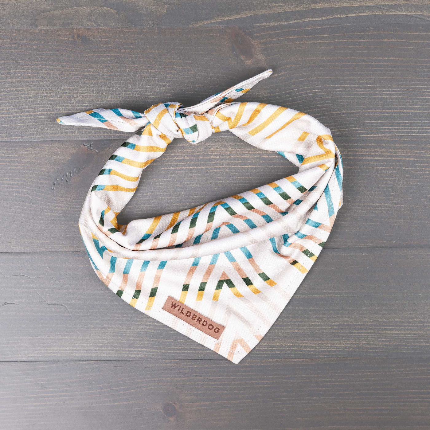 A cream bandana with vibrant diagonal stripes in blue, yellow, and green is folded and tied in a knot. It is displayed on a wooden surface. The brand "Wilderdog" is stitched in brown on a small tag attached to the bandana—perfect for your furry friend on the trail.