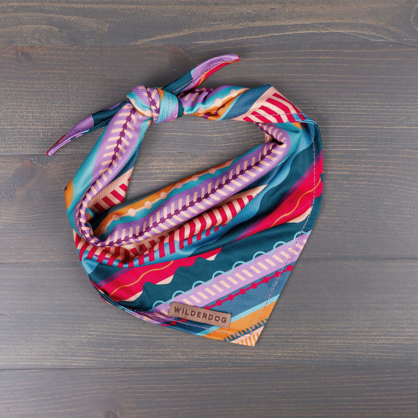 The Dawn Bandana, showcasing a vibrant geometric pattern in shades of teal, pink, purple, and coral, lies flat on a wooden surface. This colorful cooling bandana features a knot for secure fastening and is made from poly-blend mesh material for durability on any dog trail. The brand name "Wilderdog" is visible on a small tag.