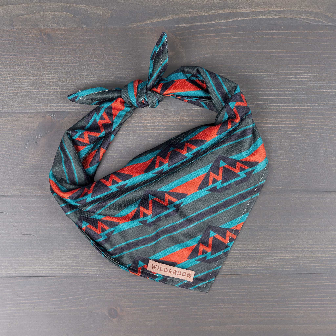 A colorful dog bandana, named Olive Bandana, featuring an abstract mountain pattern in teal, red, and black. The bandana is tied at the top and displayed on a dark wooden surface. A small tag near the bottom edge reads "WILDERDOG". Ideal for trail accessories, this stylish piece also functions as a cooling bandana for your pet.