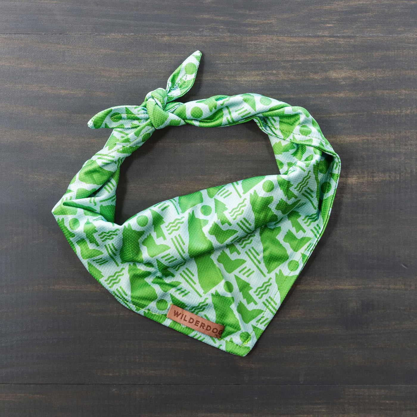 A green bandana with a white geometric pattern featuring various abstract shapes lies flat on a dark wooden surface. The cooling Pet Network Bandana has a small leather tag with the text "WILDERDOG" stitched on one corner, making it an ideal addition to your fall dog gear collection.