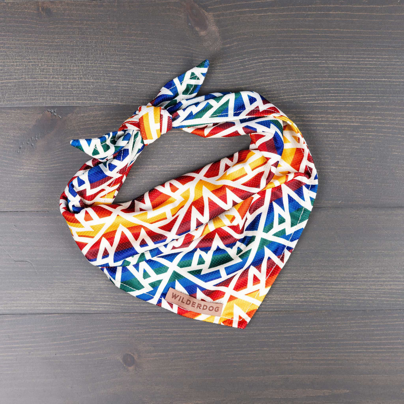 The Rainbow Bandana, with its vivid geometric pattern, rests on a dark wooden surface. This lightweight poly-blend fabric showcases a spectrum of colors—red, blue, green, yellow, and white. A "Wilderdog" label is sewn onto it and the fabric is tied in a knot on one side—ideal gear for your canine companion on the trail.