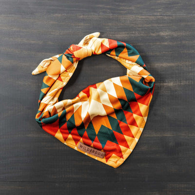 The Russet Bandana, a colorful and lightweight poly-blend mesh dog accessory featuring geometric patterns in orange, yellow, green, and black, is displayed on a dark wooden surface. Tied in a knot at the top, it's ideal for fall cooling adventures and summit selfies. A small label reads "Wilderdog.