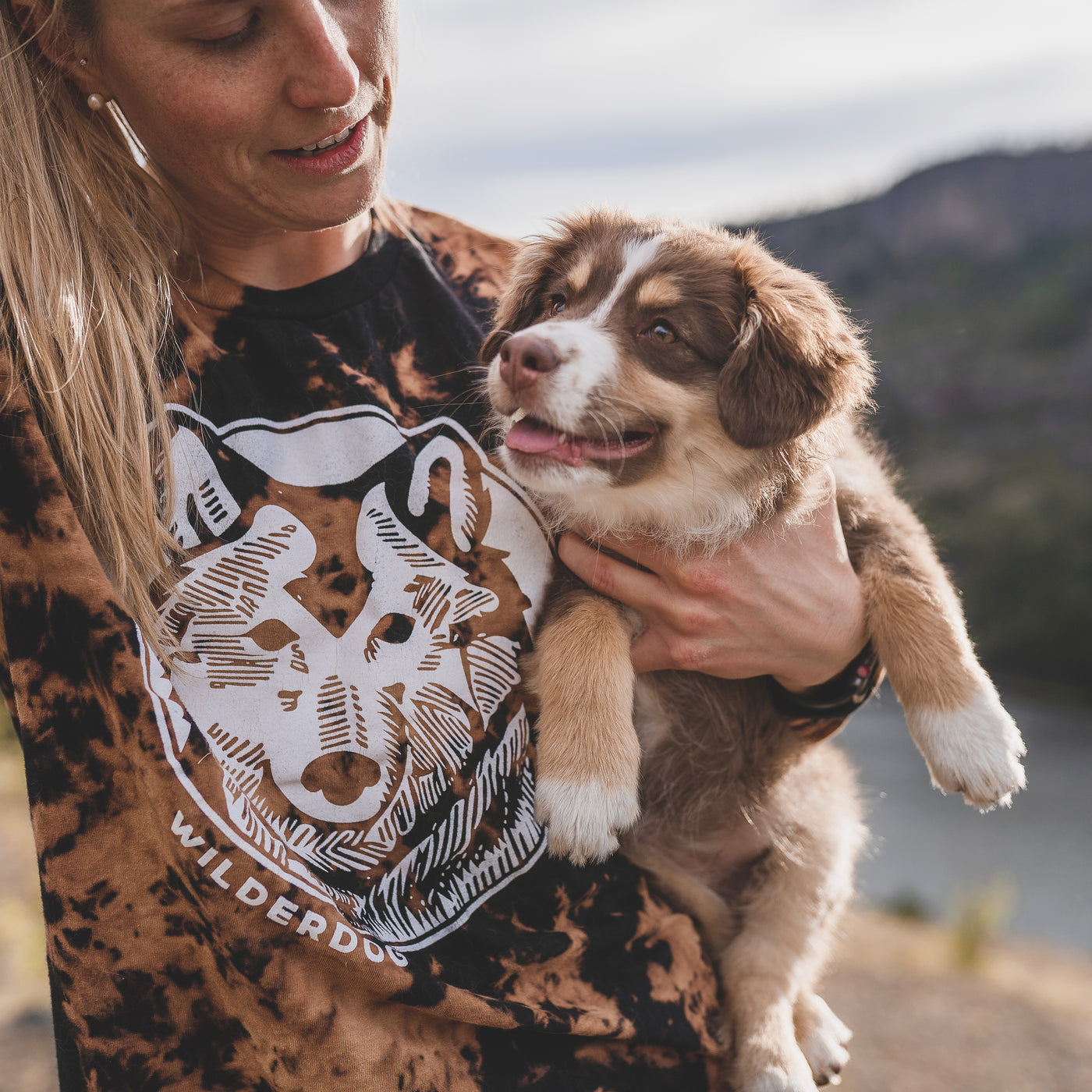 A person holding a small, fluffy, brown and white puppy outdoors. The person is wearing a Reverse Dye Long Sleeve Wolf Shirt with a graphic of a dog's face and the word "Wilderdog." The background features a blurred natural landscape with trees and hills.