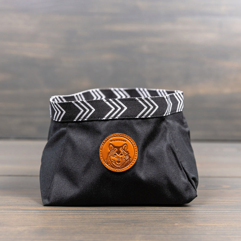 A small, black fabric Backpacking Bowl with a water-resistant herringbone-patterned rim and a leather patch featuring a dog's face rests on a wooden surface. The bowl is open, showcasing its cylindrical shape—perfect for your backpacking adventures.