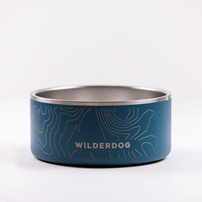 A Stainless Steel Dog Bowl that is BPA-free, showcasing a blue exterior adorned with a topographic map design and the WILDERDOG brand name printed on the side. The interior of the bowl is metallic silver, and it sits on a plain white surface with a minimalistic design.