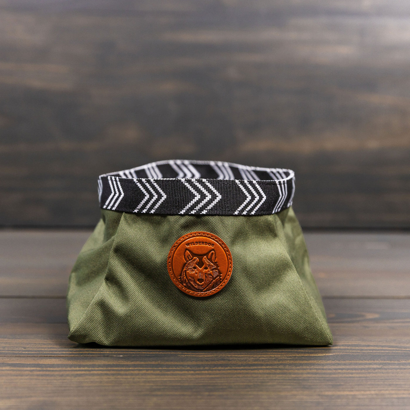 A Backpacking Bowl, featuring a collapsible green design with a black and white patterned rim, sits on a wooden surface. A brown patch with a dog logo is stitched to the front of this handy travel pet accessory, making it perfect for backpacking or any adventure.