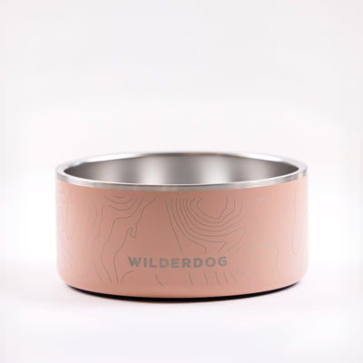 The Stainless Steel Dog Bowl is a robust and stylish option for your pet, featuring a charming light pink exterior adorned with delicate topographic lines. The side of the bowl prominently showcases the "WILDERDOG" brand name in bold capital letters. The background is set against a simple, light gray backdrop.