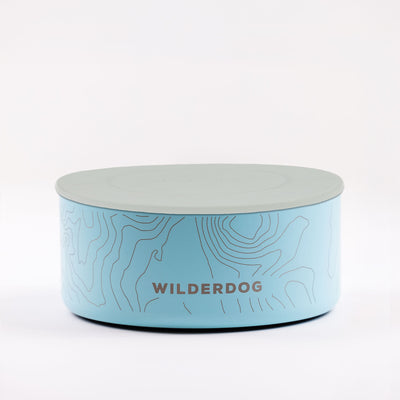 A light blue stainless steel dog bowl with a grey lid, featuring a subtle topographic map design. The text "WILDERDOG" is prominently displayed on the side in dark blue font. Made from BPA-free materials, it’s perfect as a stylish and functional accessory for your dog. The background is plain white.