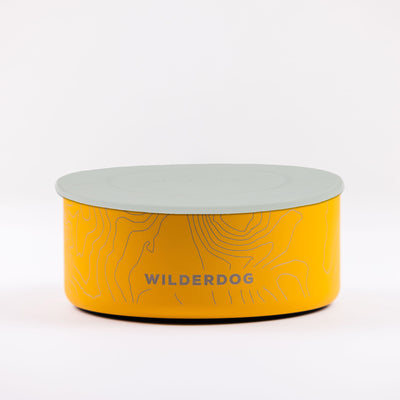 A round, yellow Stainless Steel Dog Bowl, featuring topographic design details and BPA-free construction, is displayed against a plain white background. The bowl bears the "WILDERDOG" logo on its side and comes with a light gray lid.