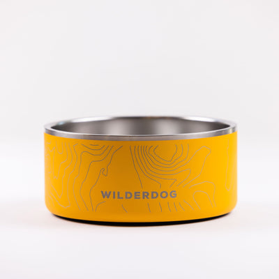 A BPA-free yellow Stainless Steel Dog Bowl featuring a topographic map design and the word "WILDERDOG" printed on the side. The interior of the bowl is silver-toned, and it sits on a plain white surface.