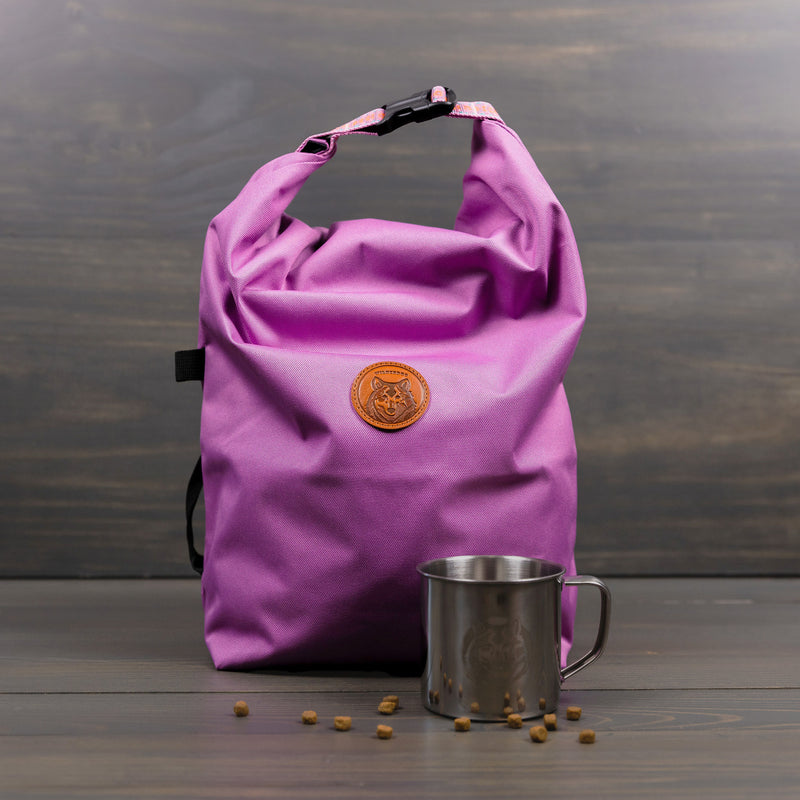 A "Doggie Bag" roll-top bag with a brown circular logo is placed on a wooden surface. In front of it sits a stainless steel mug, accompanied by small brown pellets scattered nearby, ideal for outdoor adventures. The dark gray wooden background enhances the scene&