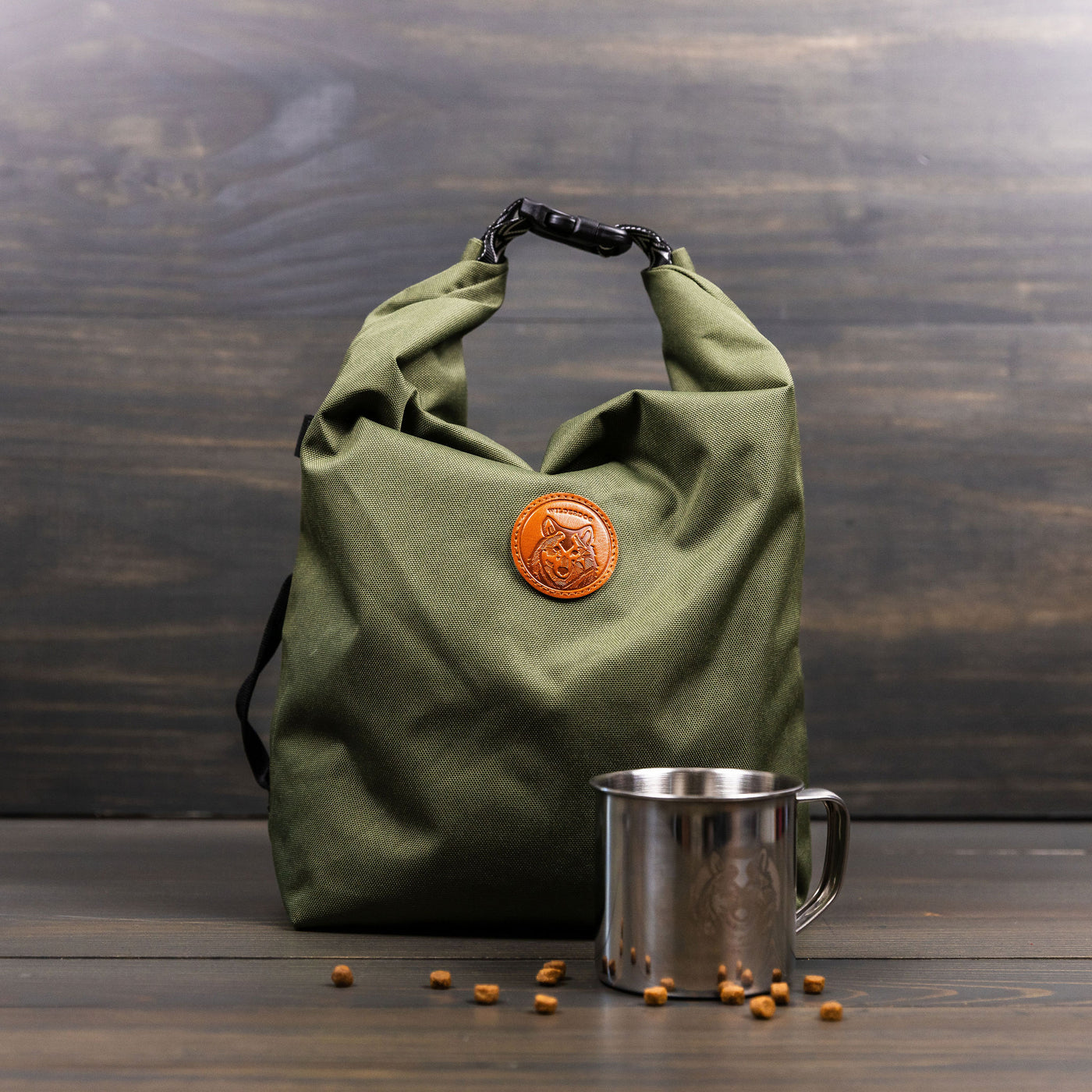 A green, roll-top Doggie Bag featuring a brown leather logo patch displayed on a wooden surface, next to a stainless steel camping mug and scattered small wooden beads. The background showcases a dark-toned wooden paneled wall. This versatile bag also doubles as a portable kibble carrier for your adventures.