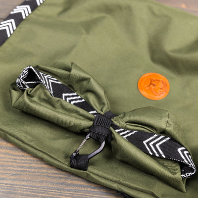 A close-up image of a green fabric Backpacking Bowl with a black and white zigzag strap and a small carabiner, perfect for attaching to your gear. The bowl is made from food-grade materials. It features a brown circular leather patch with an embossed logo and appears to be lying on a wooden surface.