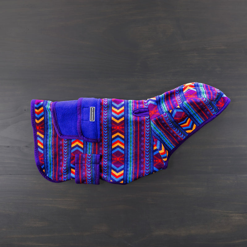 The Big Sky Fleece Jacket, designed by Wilderdog and adorned with vibrant geometric patterns in blue, red, purple, and orange hues, is displayed flat on a dark wooden surface. This jacket features a hood and a Velcro strap for secure fastening. Enjoy peace of mind with our lifetime warranty.