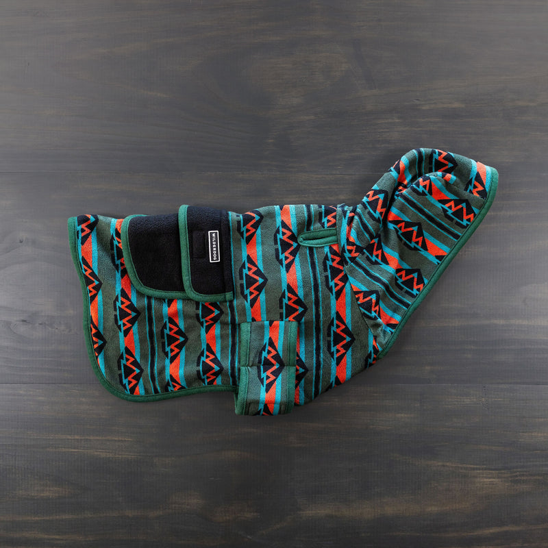 The Olive Fleece Jacket features a geometric pattern in teal, orange, red, and black. This stylish dog coat includes a black strap with Velcro for secure chest fastening and a hood. Laid flat on a dark wooden surface, the jacket's premium quality is highlighted, further supported by the Wilderdog Lifetime Warranty.