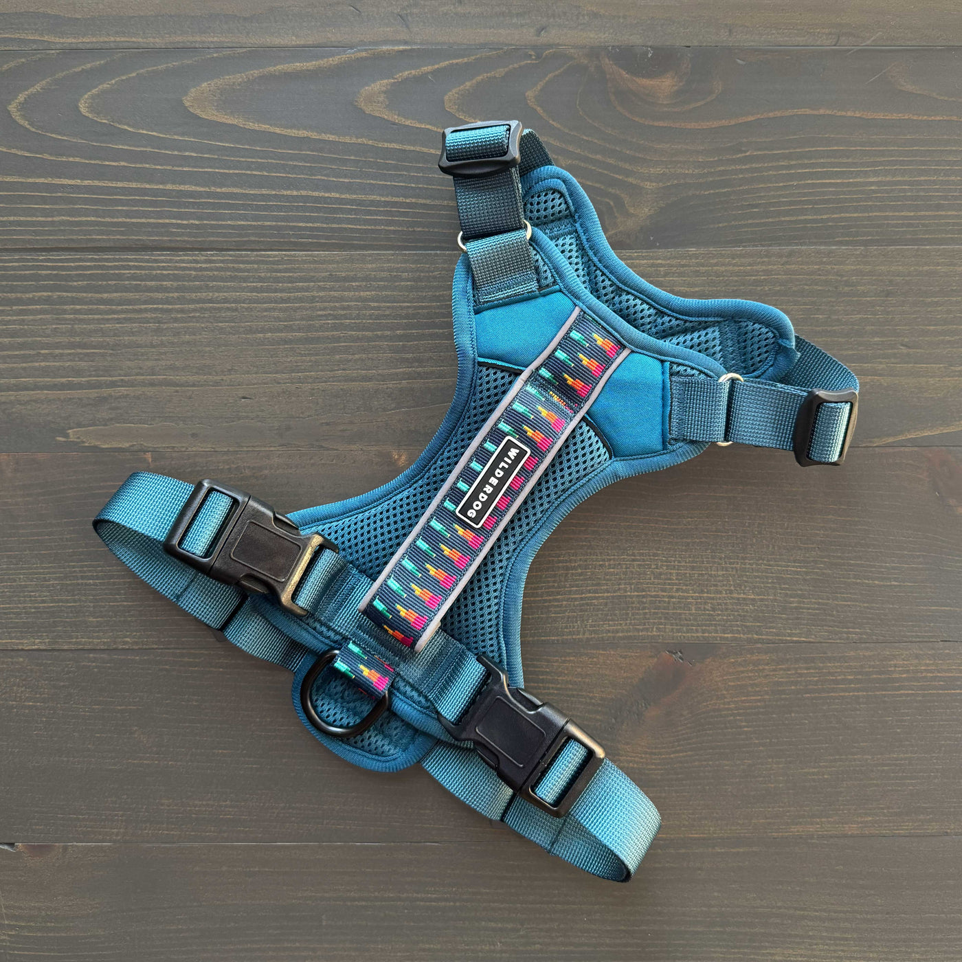 A teal-colored Dog Harness with black plastic buckles and adjustable straps lies flat on a wooden surface. The lightweight mesh fabric showcases a decorative multicolored stripe along the chest section and reflective strips for added safety, while a metal D-ring provides secure leash attachment.