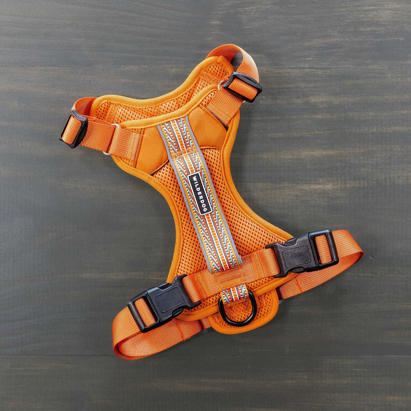 An orange Dog Harness with reflective material is placed on a dark wooden surface. It features a lightweight mesh design, adjustable straps, sturdy buckles, and a metal ring for leash attachment.