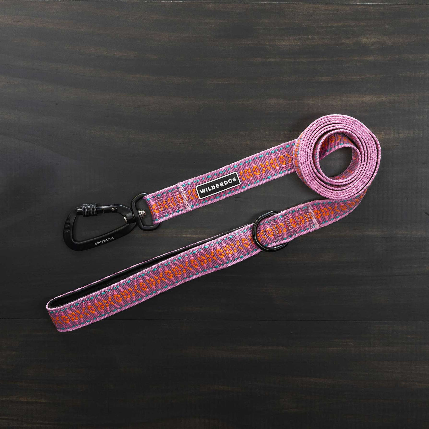 The Lilac Ultralight Leash, adorned with a vibrant pattern of pink, orange, and purple, is fully extended on a dark wooden surface. It features a black metal clasp and an adjustable padded handle. Part of the Ultralight Collection from Wilderdog, it includes a tag that reads "Wilderdog.