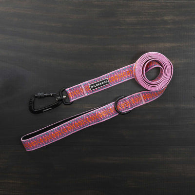 The Lilac Ultralight Leash, adorned with a vibrant pattern of pink, orange, and purple, is fully extended on a dark wooden surface. It features a black metal clasp and an adjustable padded handle. Part of the Ultralight Collection from Wilderdog, it includes a tag that reads "Wilderdog.