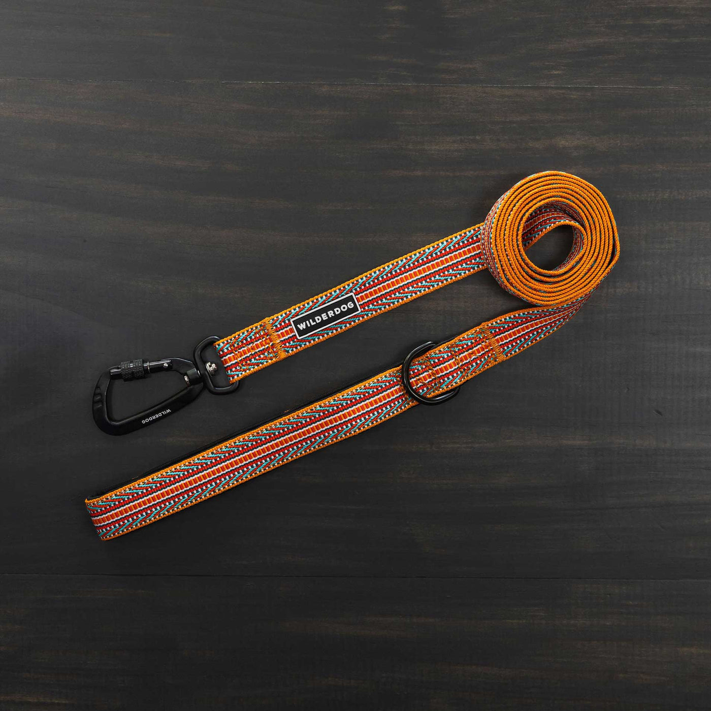 The Sienna Ultralight Leash, a vibrant and durable accessory made from strong nylon webbing, is laid out on a dark wood surface. This stylish leash features orange, yellow, and red patterns woven into its design. It includes a swiveling carabiner attachment for easy use, a metal D-ring for added functionality, and proudly displays the "Wilderdog" label.