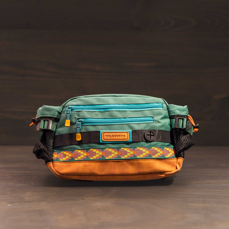 A vibrant Mega Hip Pack featuring a green top, a blue front zipper, and an orange-brown bottom. It showcases a multicolored geometric pattern strip and includes a "WILDBROOS" label tag. The bag comes with an adjustable strap and offers a 3-liter capacity, all set against a dark background.