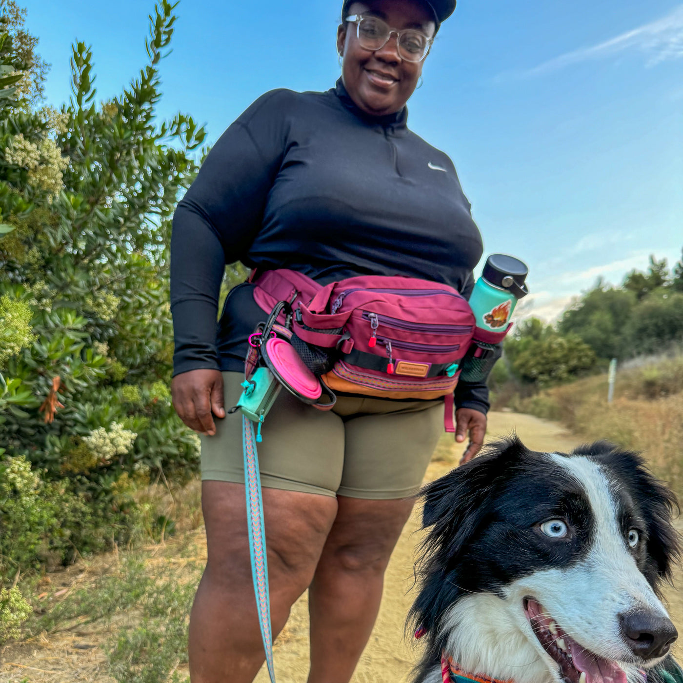 A person in a black long-sleeve shirt, beige shorts, and a Mega Hip Pack with an adjustable strap stands outdoors with a dog. The person wears glasses and a cap, and holds a water bottle. The dog has a black-and-white coat with blue eyes. They appear to be on a nature trail.