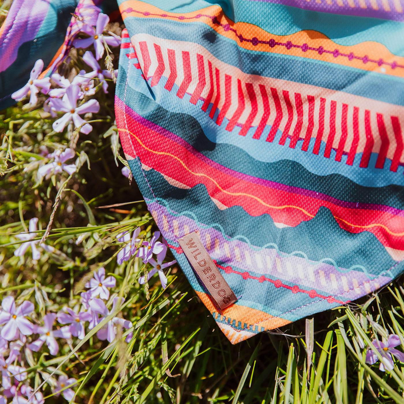 The Dawn Bandana, showcasing a vibrant geometric pattern in shades of blue, red, and orange, is draped over a backdrop of purple flowers and green grass. Made from poly-blend mesh material, the bandana features a small tag that reads "Wilderdog." The scene is brightly lit, emphasizing the lively colors and textures.