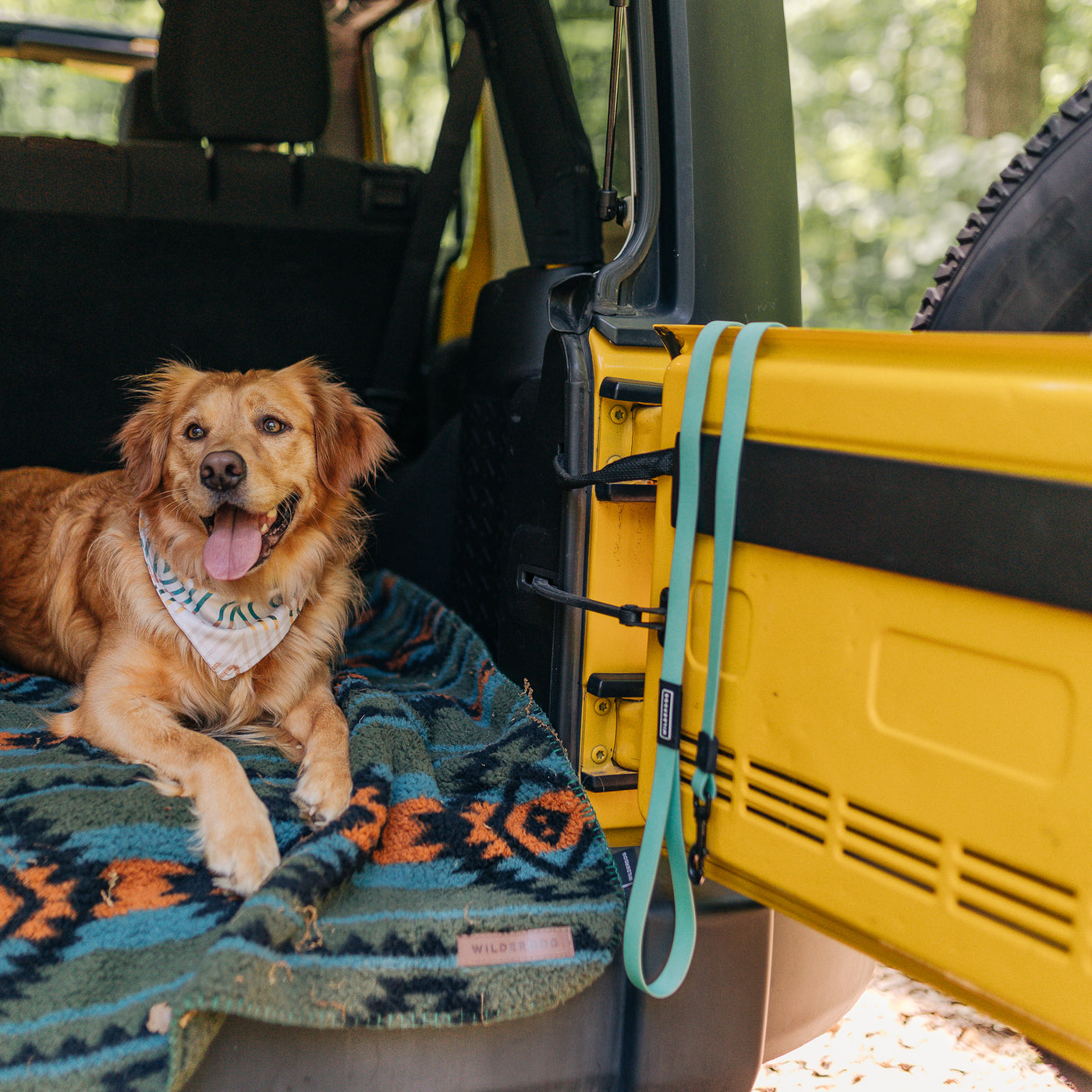 A dog with golden fur, wearing a cream poly-blend mesh bandana, lies comfortably on a colorful blanket in the open trunk of a yellow vehicle, with a leash hanging on the door. The background shows a forest setting with green foliage.