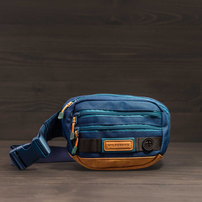 A small Utility Pack in blue and brown, crafted from vegan material with multiple zippered compartments, is set against a dark wooden background. The bag proudly displays a "Wilderdog" label and includes a sturdy adjustable strap, making it ideal for everyday adventures.