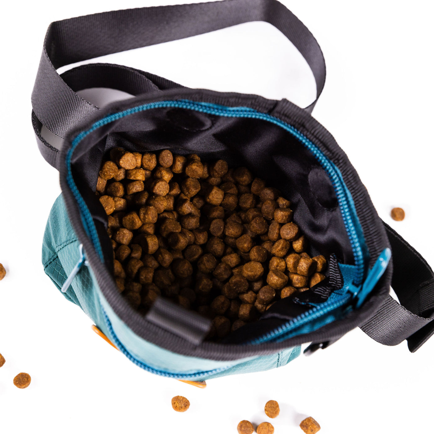 A top-down view of an open black and turquoise Dog Training Treat Pouch filled with brown spherical pet food pellets displays a few scattered pellets on the white surface. The pouch has an adjustable waist strap, a turquoise zipper, and a magnetic closure.