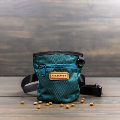 A teal Dog Training Treat Pouch with black trim and an adjustable waist strap is displayed on a wooden surface. The pouch features a small front pocket with a blue zipper, magnetic and zipper closures, and a tan patch that reads "Wilderdog." A few dog treats are scattered around the pouch.