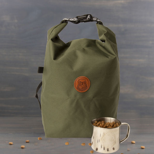 A '24 Olive Doggie Bag with a circular brown leather patch sits on a gray surface. Next to it, a stainless steel mug is filled with kibble and some scattered pieces. The background displays a gray, textured wall.