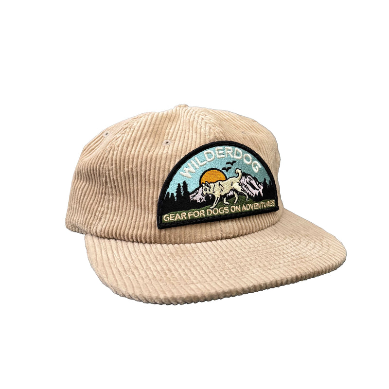 The Wilderdog Cord Hat is a beige 100% cotton cap with a vibrant dog, sunburst, and trees patch. It features "Wilderdog" and "Gear for Dogs on Adventures" embroidery, plus an adjustable clasp for the perfect fit.