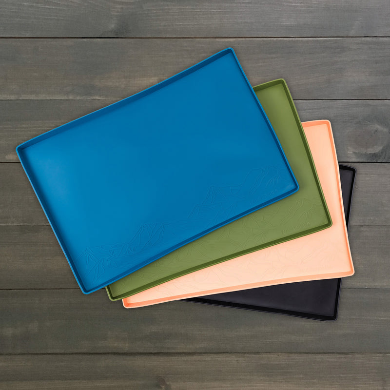 Four rectangular Silicone Placemats in blue, green, peach, and black are stacked on a wooden surface. Made from food-grade silicone with a subtle mountain design on one side, the non-slip material ensures stability and elegance in any setting.